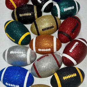 May include: A collection of glitter-covered football ornaments in various colors, including red, green, gold, silver, and blue. Each football has white or yellow stripes and stitching details, creating a festive and decorative appearance.