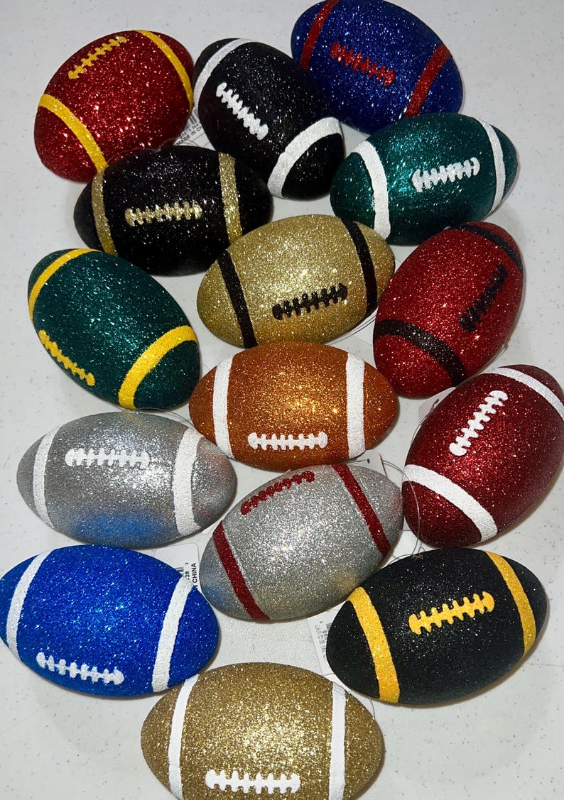 May include: A collection of glitter-covered football ornaments in various colors, including red, green, gold, silver, and blue. Each football has white or yellow stripes and stitching details, creating a festive and decorative appearance.