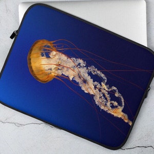 Op de afbeelding: Een laptophoes met een levendige blauwe achtergrond waarop een grote, doorschijnende kwal met een goud-oranje klok en lange, hangende tentakels is afgebeeld.