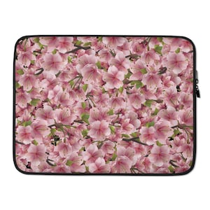 Puede incluir: Funda para portátil con un diseño floral de flores de cerezo rosas. El diseño incluye hojas verdes y ramas marrones sobre un fondo negro. La funda tiene una cremallera y un ribete negros.