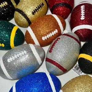 May include: A collection of glitter-covered football ornaments in various colors, including gold, silver, red, blue, and green. Each ornament features white or contrasting-color stripes and stitching details, resembling miniature American footballs.