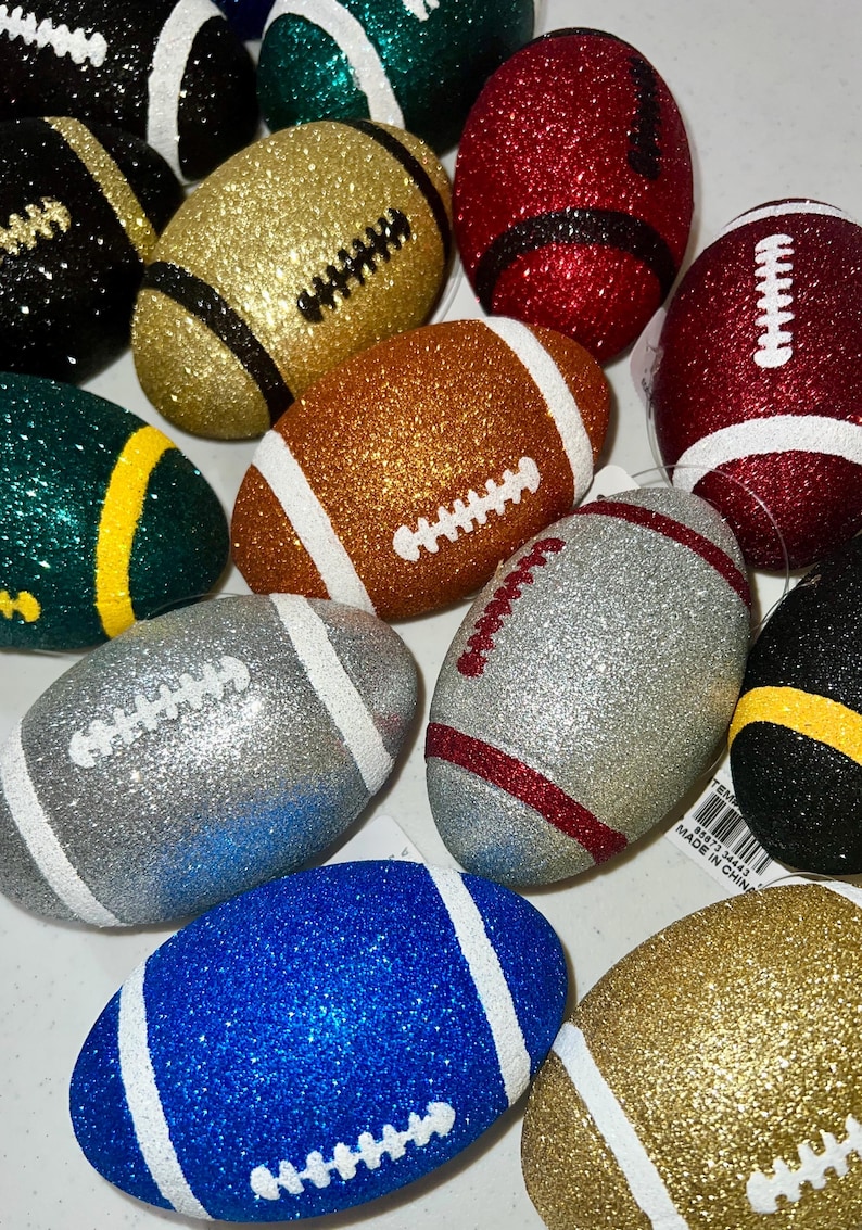 May include: A collection of glitter-covered football ornaments in various colors, including gold, silver, red, blue, and green. Each ornament features white or contrasting-color stripes and stitching details, resembling miniature American footballs.