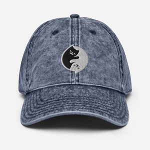 May include: A blue denim baseball cap with a black and white yin yang symbol featuring two cats facing each other. The symbol is embroidered on the front of the cap.