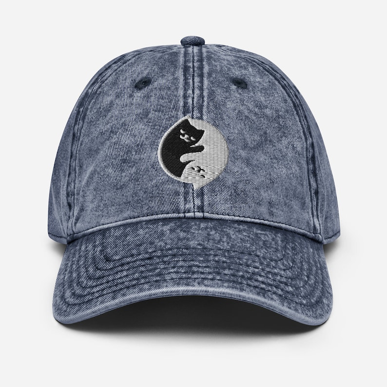 May include: A blue denim baseball cap with a black and white yin yang symbol featuring two cats facing each other. The symbol is embroidered on the front of the cap.