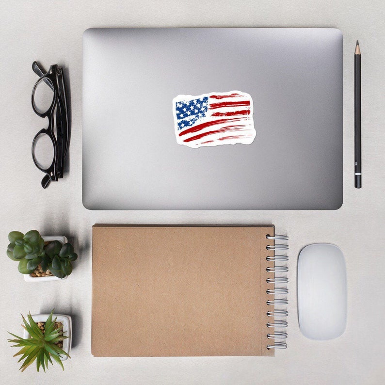 May include: A distressed American flag sticker on a silver laptop computer. The sticker is white with red and blue stripes and white stars.