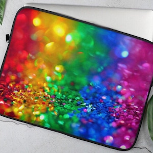 Op de afbeelding: Een laptophoes in regenboogkleuren met een glinsterende textuur. De hoes heeft een wazige achtergrond met kleurrijke lichten.