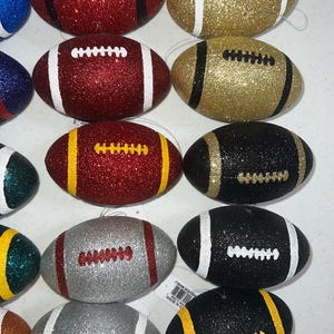May include: Glitter-covered football ornaments in various colors, including red, gold, black, and silver. Each ornament features white or yellow stripes and laces, perfect for holiday decorations or sports-themed crafts.