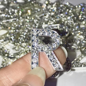 Peut inclure: Une lettre R argentée recouverte de strass transparents. La lettre est tenue dans une main.