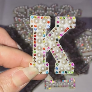 May include: A white letter K covered in white pearls and colourful rhinestones.