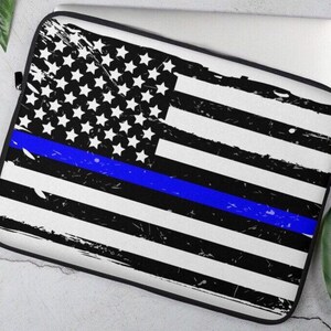 Puede incluir: Una funda para portátil con un diseño de bandera estadounidense desgastado con una fina línea azul en el medio. La bandera es negra y blanca con una línea azul.