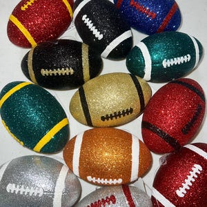 May include: A collection of glitter-covered football ornaments in various colors, including red, black, gold, green, blue, silver, and orange. Each ornament has white or yellow stripes and stitching details, resembling miniature American footballs.