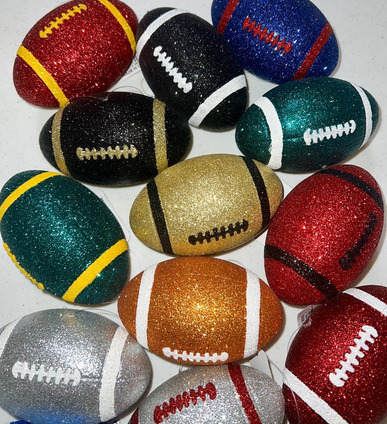 May include: A collection of glitter-covered football ornaments in various colors, including red, black, gold, green, blue, silver, and orange. Each ornament has white or yellow stripes and stitching details, resembling miniature American footballs.