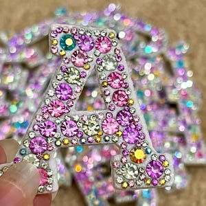 Puede incluir: Primer plano de una brillante letra "A" adornada con pedrería colorida en tonos rosa, morado, amarillo y verde. La letra es blanca con un acabado brillante, creando un efecto deslumbrante. Otras letras son visibles en el fondo.