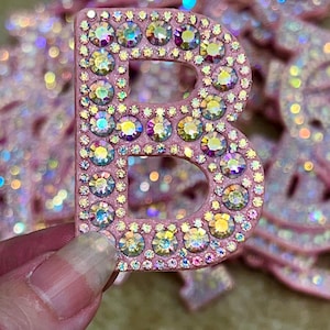 Puede incluir: Una letra "B" rosa incrustada con pedrería iridiscente. La letra se sostiene en una mano, con otras letras similares visibles en el fondo. Las piedras reflejan varios colores, creando un efecto brillante.