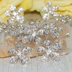May include: Silver flower-shaped embellishments with clear rhinestones. Each flower has five points and is approximately 1 inch in diameter.