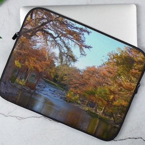 Könnte beinhalten: Eine Laptop-Hülle mit einem Landschaftsbild eines Flusses, der durch einen Wald mit Herbstlaub in Orange-, Gelb- und Grüntönen fließt.