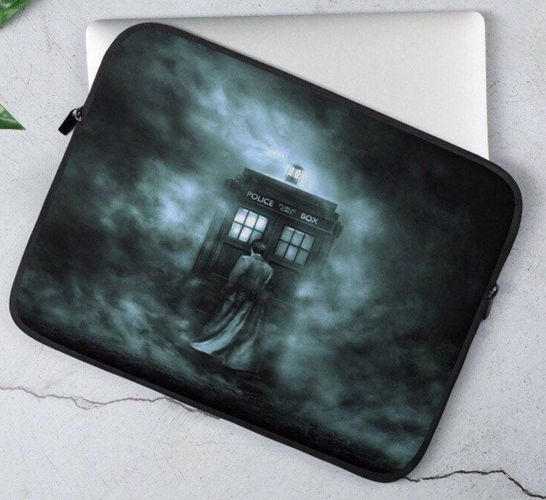 May include: Black laptop sleeve featuring a TARDIS police box design in a misty, dark blue and gray scene. The words "POLICE BOX" are visible on the top of the box. The sleeve has a black zipper closure and is resting on a silver laptop.