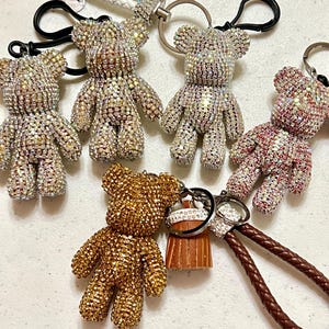 Puede incluir: Varios llaveros de osos de peluche cubiertos de diamantes de imitación, en plata, oro y rosa. Cada oso tiene un cierre de metal y una correa en colores como blanco y marrón. Los llaveros son decorativos.