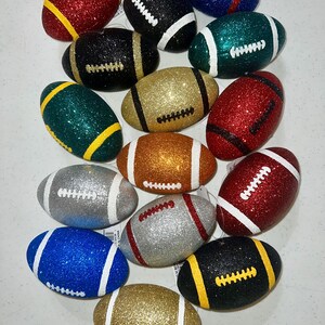 May include: A collection of glitter-covered football ornaments in various colors, including red, black, gold, green, silver, blue, and orange. Each ornament features white or yellow stitching details and stripes, creating a festive and decorative appearance.
