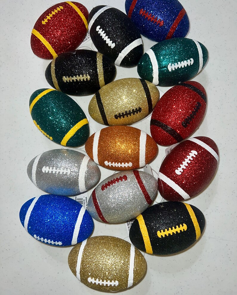 May include: A collection of glitter-covered football ornaments in various colors, including red, black, gold, green, silver, blue, and orange. Each ornament features white or yellow stitching details and stripes, creating a festive and decorative appearance.
