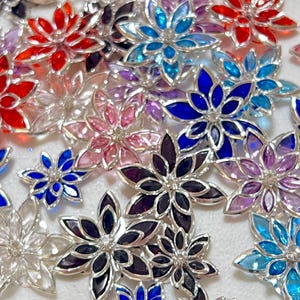 May include: A collection of flower-shaped embellishments in various colors, including red, blue, pink, purple, and clear. Each flower has a silver-colored metal frame and a central crystal. The flowers are scattered across a white surface.