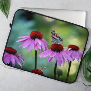 May include: A butterfly with black, white, and orange wings sits on a pink coneflower. The flower has a red center and is surrounded by green leaves.