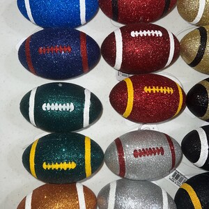 May include: A collection of glittery football ornaments in various colors, including blue, red, green, gold, silver, and brown. Each ornament features white or yellow stripes and laces. These decorative ornaments are designed for festive occasions.