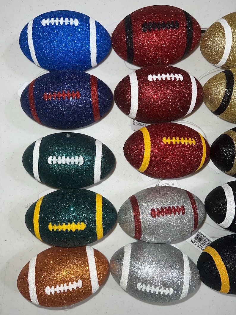 May include: A collection of glittery football ornaments in various colors, including blue, red, green, gold, silver, and brown. Each ornament features white or yellow stripes and laces. These decorative ornaments are designed for festive occasions.