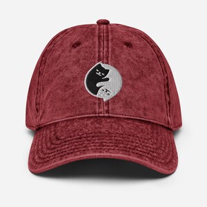 May include: A red vintage washed cotton baseball cap with an embroidered yin yang symbol featuring two black cats facing each other.