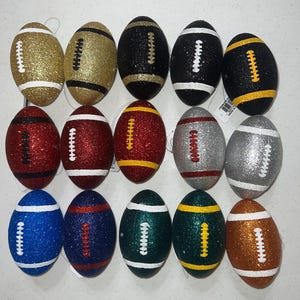 May include: A collection of fifteen glitter-covered football ornaments in various colors. The ornaments include gold, black, red, silver, blue, green, and brown, each with white or yellow stitching details. These decorative footballs are ideal for sports-themed decor.