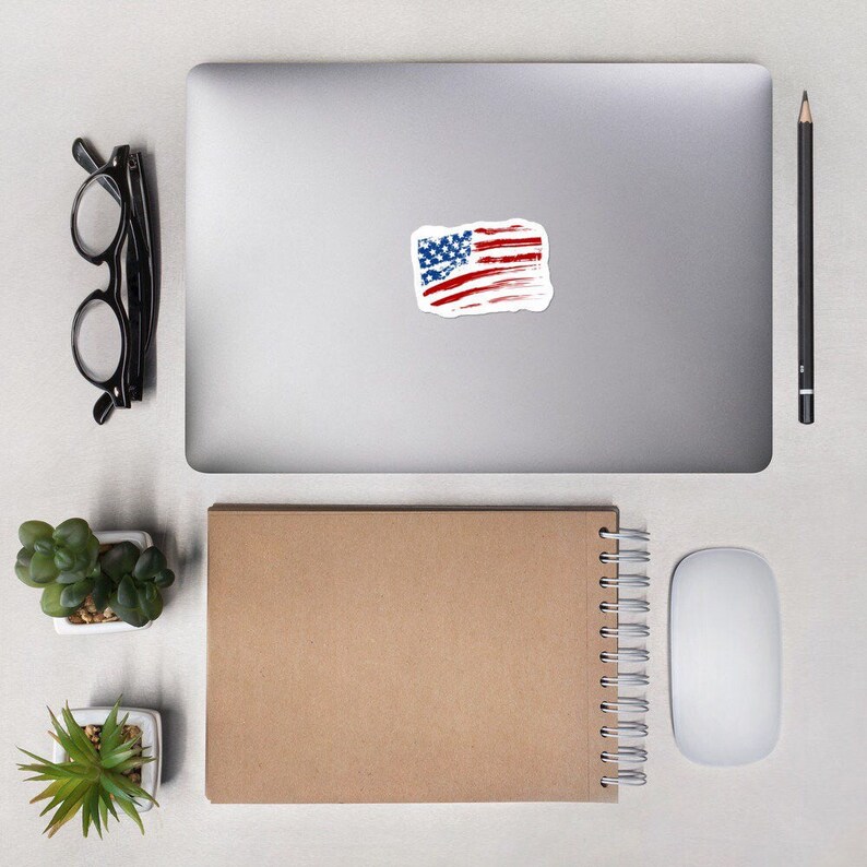 May include: A distressed American flag sticker on a silver laptop computer. The sticker is a white background with red and blue stripes and white stars.