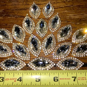 May include: A collection of 18 clear crystal rhinestones with gold settings. The rhinestones are arranged in a triangular shape and are approximately 2.5 cm in length. The rhinestones are measured with a yellow tape measure.