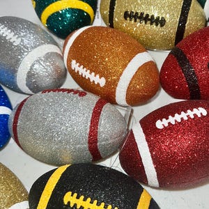 May include: A collection of glitter-covered football ornaments in various colors, including gold, silver, red, green, and black. Each ornament features white or yellow stitching and stripes, creating a festive and decorative appearance.