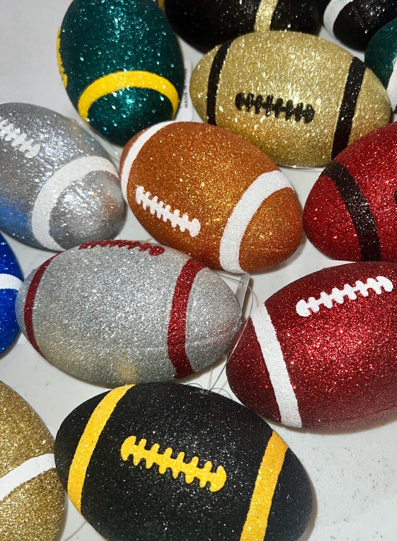 May include: A collection of glitter-covered football ornaments in various colors, including gold, silver, red, green, and black. Each ornament features white or yellow stitching and stripes, creating a festive and decorative appearance.