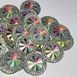 May include: A group of iridescent rainbow-colored round rhinestones with a silver trim. The rhinestones have a faceted design and are arranged in a pile.