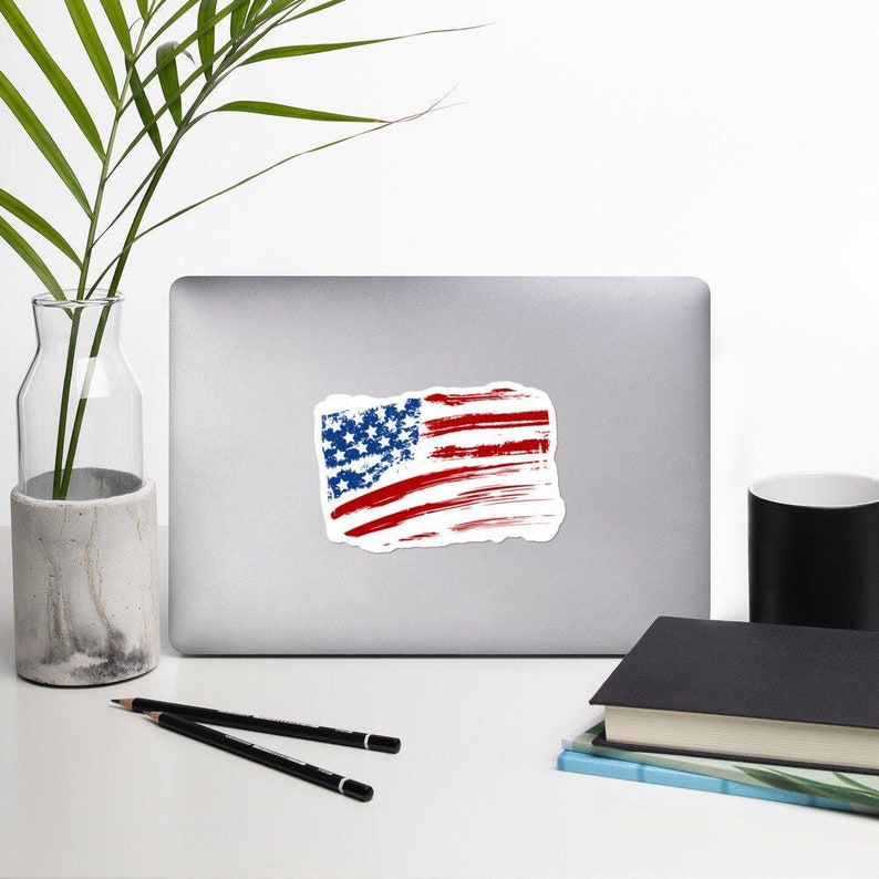 May include: A distressed American flag sticker with red and white stripes and blue stars. The flag is slightly faded and has a worn look.