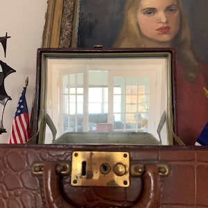 May include: A brown leather suitcase with a brass lock and handle. The suitcase is open and a mirror inside reflects a room with windows. A painting of a woman with blonde hair is in the background.