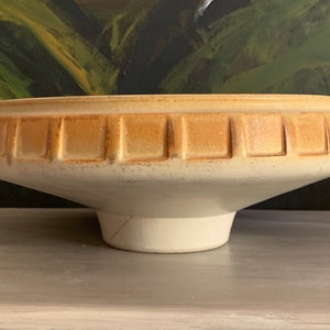 May include: A large, white ceramic bowl with a wide, flared rim and a raised, geometric design in a light brown glaze. The bowl sits on a wooden surface.