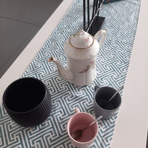 Könnte beinhalten: Eine weiße Teekanne mit Blumendekor steht auf einem türkisfarbenen Tischläufer mit geometrischem Muster. Auf dem Tischläufer stehen drei Tassen, eine schwarze, eine rosa und eine graue.