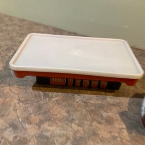 May include: A white plastic container with a red base and a clear lid. The container has multiple compartments and is sitting on a brown surface.