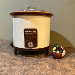 以下が含まれることがあります： クリームと茶色のヴィンテージHamilton Beach「Simmer-on」スロークッカー。透明な蓋と「オフ」、「ロー」、「ハイ」の設定があるコントロールパネルがあります。装飾用の陶器製アヒルの置物がスロークッカーの隣にあります。