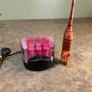 May include: A hair roller set with pink rollers and a black base, next to a carved wooden orange and black tiger figurine. The set is plugged into an electrical outlet.