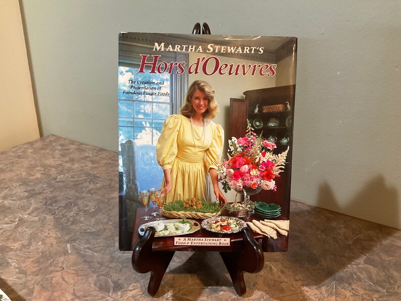 May include: A cookbook titled "Martha Stewart's Hors d'Oeuvres: The Creation and Presentation of Fabulous Finger Foods" with a photo of Martha Stewart on the cover. The cover features a yellow dress, a floral arrangement, and a table setting with food.