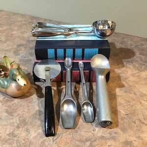May include: A set of kitchen utensils including a pizza cutter, two measuring spoons, a small ice cream scoop, and a larger ice cream scoop with a handle. The utensils are arranged on top of two blue books.