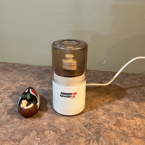 Può includere: Un tritatutto elettrico bianco con un coperchio in plastica trasparente. Il tritatutto ha la scritta "Herbie Jr. Grain" stampata sul lato. In primo piano c'è una piccola statuetta di anatra marrone e verde.