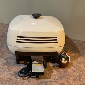 Puede incluir: Parrilla eléctrica de encimera blanca con base marrón y un pomo negro en la tapa. La parrilla tiene un panel de control con un dial y una serie de luces que indican diferentes temperaturas de cocción. El panel de control tiene el texto "Sensor-Temp Heat Control" y enumera varios alimentos y sus temperaturas correspondientes. Una pequeña figurita de pato de cerámica está en primer plano.