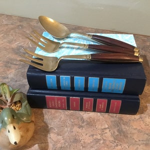 以下が含まれることがあります： スプーンとフォーク2本を含む、木製のダークハンドルが付いたヴィンテージ真鍮製カトラリーセット。青と赤の本の上に置かれています。カトラリーは、薄い青色の模様の紙の上にあります。