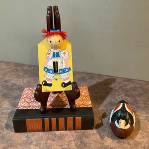 May include: A wooden light switch cover with a painted image of a girl in a blue dress and red hair. The cover is on a wooden easel and a painted wooden duck is on the table.