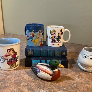 Puede incluir: Una colección de cuatro tazas de cerámica con personajes de Disney. Las tazas son blancas con diseños coloridos. Una taza presenta a Mickey y Minnie Mouse, otra presenta a Grumpy de Blancanieves, y otra presenta una variedad de personajes de Disney. La cuarta taza es blanca con un diseño azul y blanco. Las tazas están dispuestas sobre una mesa con una pila de libros y una figurita de pato de madera.