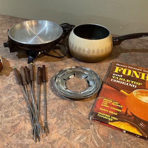 Op de afbeelding: Een vintage fondueset met een bruine en witte keramische pot, een metalen basis en een set houten vorken met handvatten. De set wordt geleverd met een kookboek getiteld "Fondue en tafelkoken" van Better Homes and Gardens.